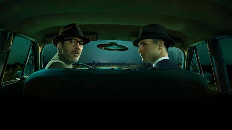 The two main characters are sitting in a car with doubtful expressions
